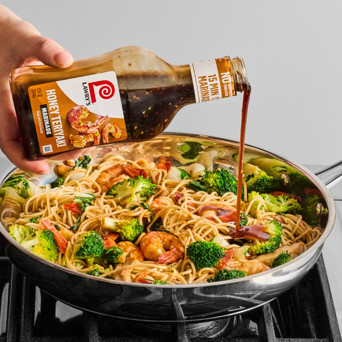 A hand pours Lawrys Honey Teriyaki Marinade over a pan of noodles shrimp and broccoli.