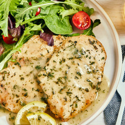 ['Two pieces of chicken breast with lemon slices and a green salad on a white plate.']