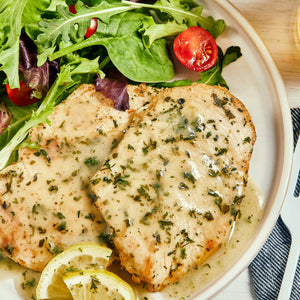 ['Two pieces of chicken breast with lemon slices and a green salad on a white plate.']