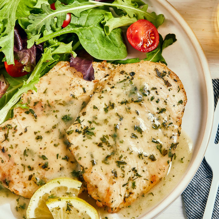 ['Two pieces of chicken breast with lemon slices and a green salad on a white plate.']