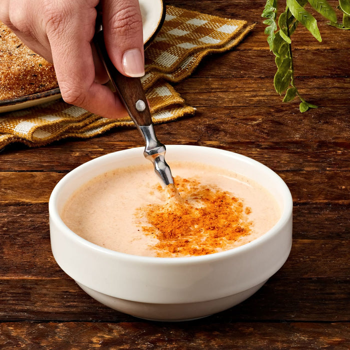 A hand stirs a bowl of creamy soup with a sprinkle of spice on a wooden table.
