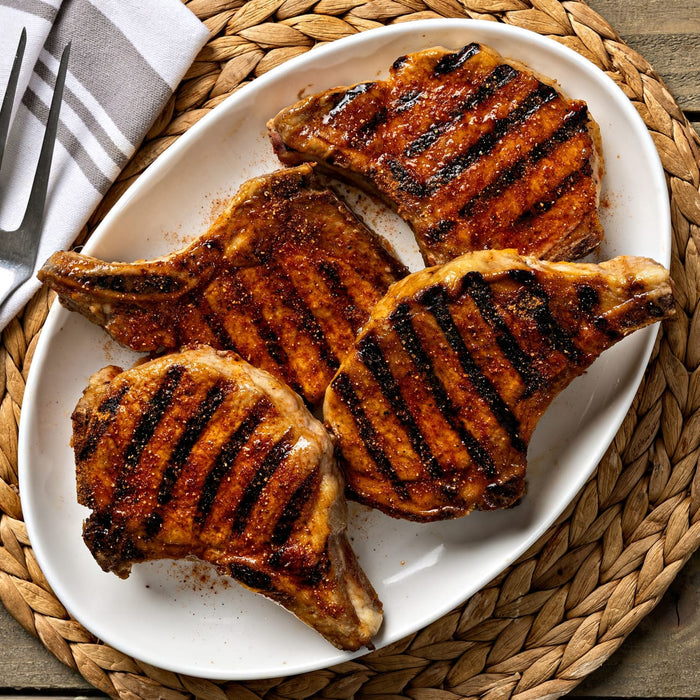 Four grilled pork chops are arranged on a white platter, garnished with spices and served with a fork and striped napkin on a woven placemat.