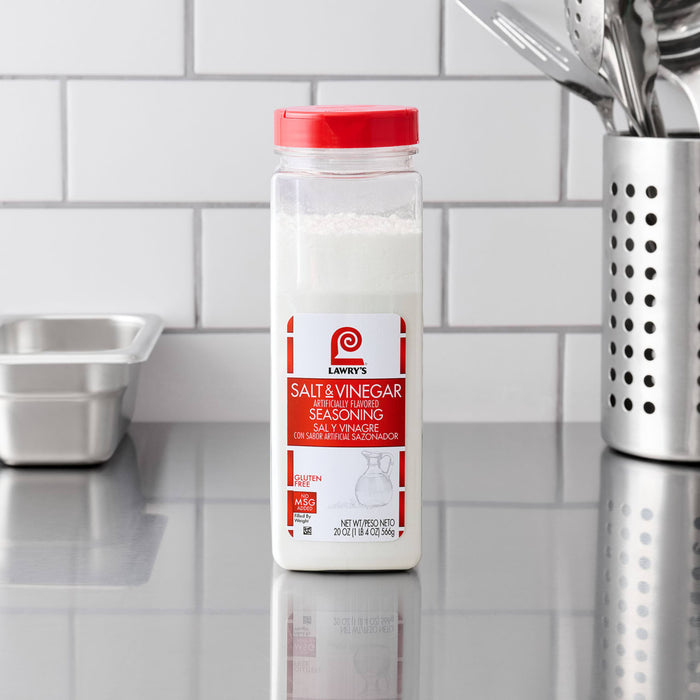 A clear shaker of Lawry'S Seasoning Salt &amp; Vinegar, 20 Oz, sits on a stainless steel countertop in a professional kitchen setting.