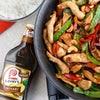 ["A bottle of Lawry's Teriyaki marinade next to a wok with chicken, cashews, snow peas, and red peppers."]