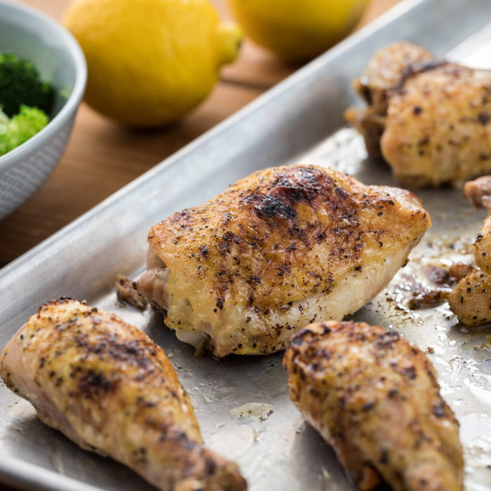 ['A close-up shot of roasted chicken pieces on a metal tray, accompanied by lemons and a bowl of broccoli.']