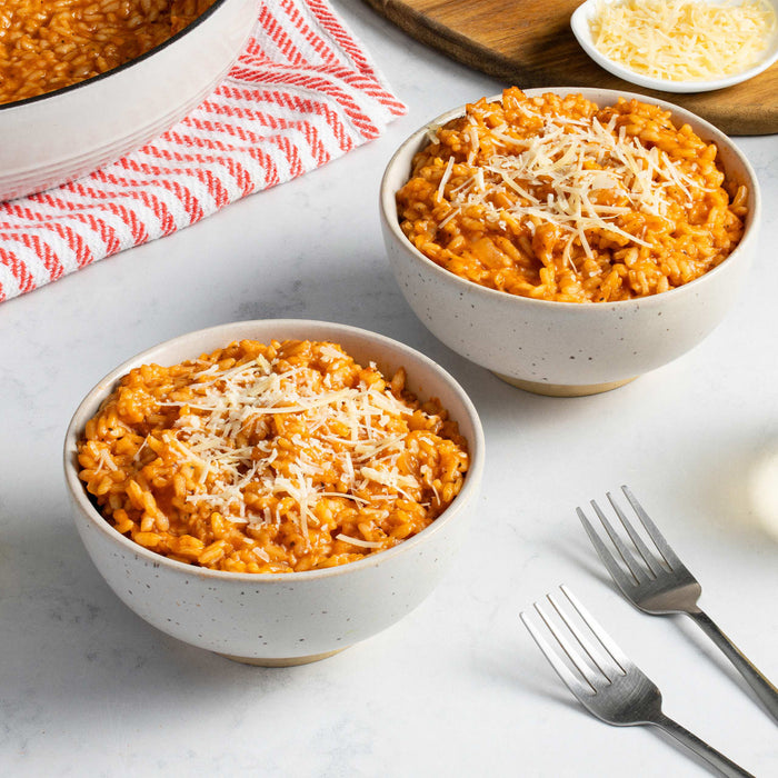 Two bowls of tomato risotto topped with shredded cheese are displayed on a white surface with forks, a pot, a red and white towel, and a small bowl of cheese.