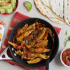 A skillet of chicken fajitas with red peppers and onions is served with tortillas, guacamole, salsa, and a lime wedge.