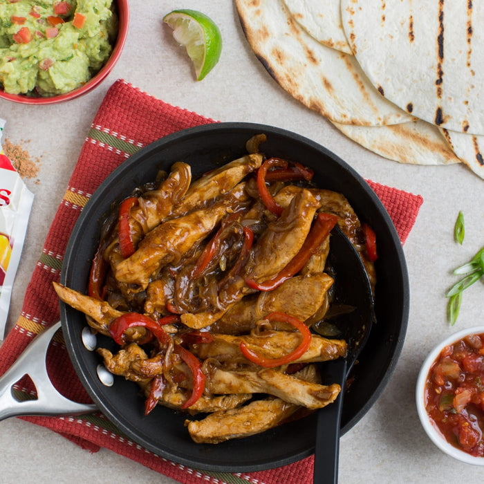 A skillet of chicken fajitas with red peppers and onions is served with tortillas, guacamole, salsa, and a lime wedge.