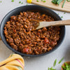 A skillet filled with chili, featuring ground beef, beans, and tomatoes, is being stirred with a wooden spoon.