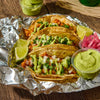 ['Three tacos topped with cilantro, onions, and a green sauce are on a bed of aluminum foil.']