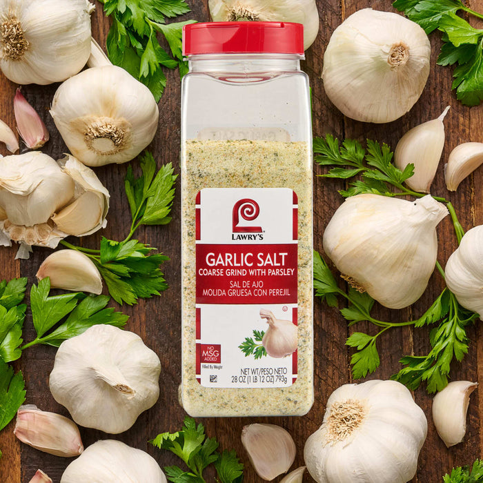 A container of Penzey's Garlic Salt is surrounded by fresh garlic bulbs and parsley.