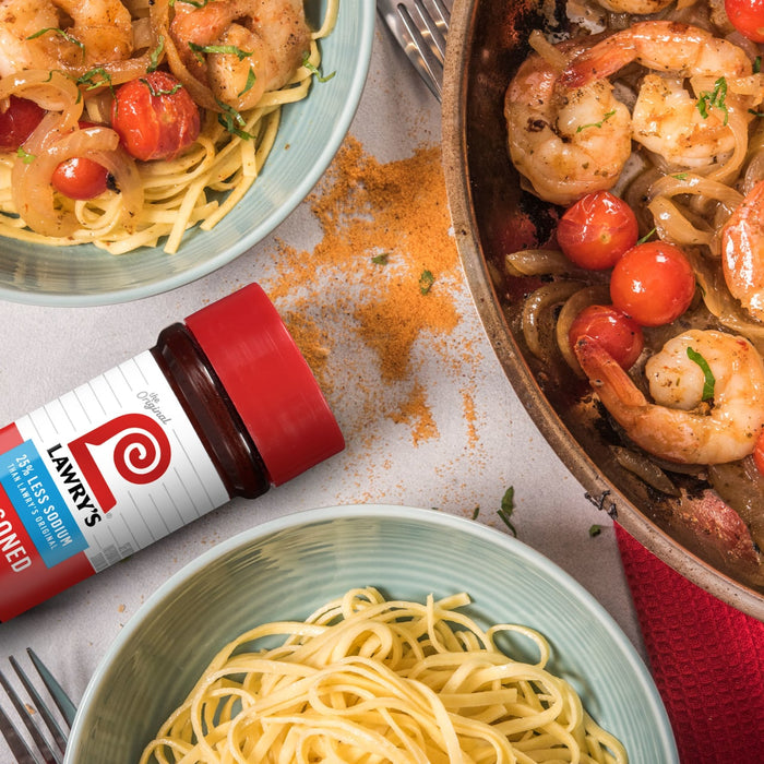 ["A bottle of Lawry's Seasoned Salt, a plate of pasta, and a pan of shrimp, tomatoes, and onions."]