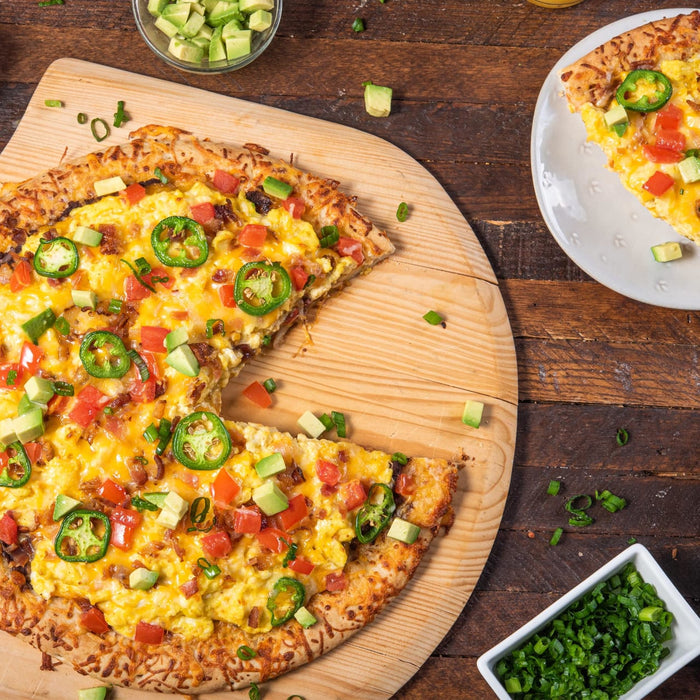 A breakfast pizza with eggs, bacon, jalapenos, tomatoes, avocado, and cheese on a wooden cutting board.
