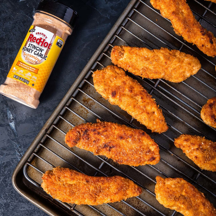 A bottle of Franks RedHot Stingin Honey Garlic Seasoning Blend next to a baking sheet with breaded chicken tenders.