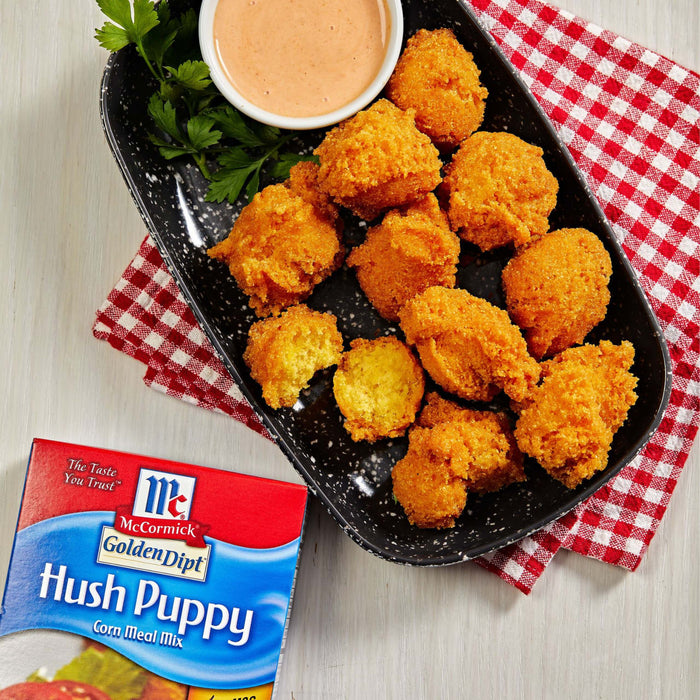 ['A plate of golden brown hush puppies with a side of dipping sauce and a box of McCormick Golden Dipt Hush Puppy Corn Meal Mix.']