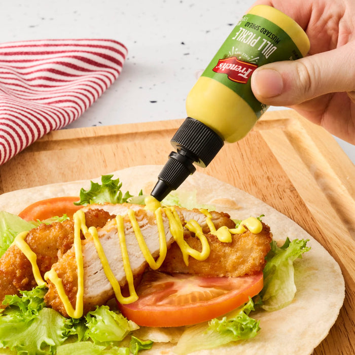 A hand squeezes French's Dill Pickle Mustard Spread onto a chicken wrap with lettuce and tomato.