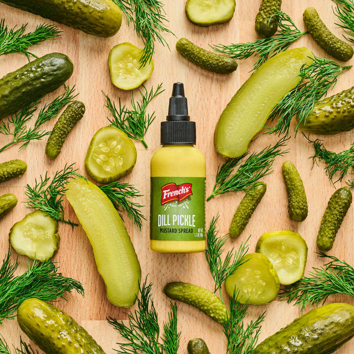 A bottle of French's Dill Pickle Mustard Spread is surrounded by pickles and dill sprigs on a wooden surface.