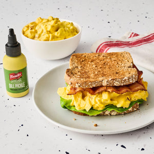 A French's Dill Pickle Mustard Spread bottle is featured next to a sandwich with egg salad, bacon, and lettuce, and a bowl of egg salad.