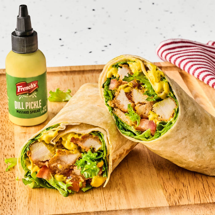 Two halves of a wrap filled with lettuce, tomatoes, chicken, and French's Dill Pickle Mustard Spread are displayed on a wooden board next to a bottle of the mustard.
