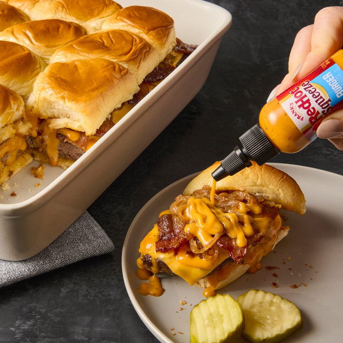 A hand squeezes Franks RedHot Buffalo Wing Sauce onto a slider with bacon cheese and caramelized onions.