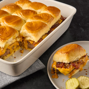 A white baking dish filled with slider buns topped with melted cheese bacon and caramelized onions with one slider on a plate with pickles.