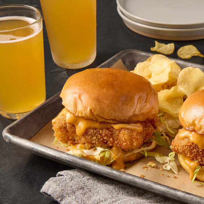 Two crispy chicken sandwiches with lettuce and sauce on a metal tray with potato chips.
