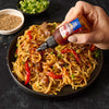 A hand is pouring Franks RedHot Korean BBQ Sauce over a bowl of noodles beef peppers and sesame seeds.