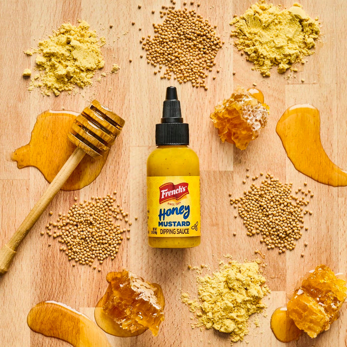 A bottle of Frenchs Honey Mustard Dipping Sauce is surrounded by honey honeycomb mustard seeds and mustard powder on a wooden surface.
