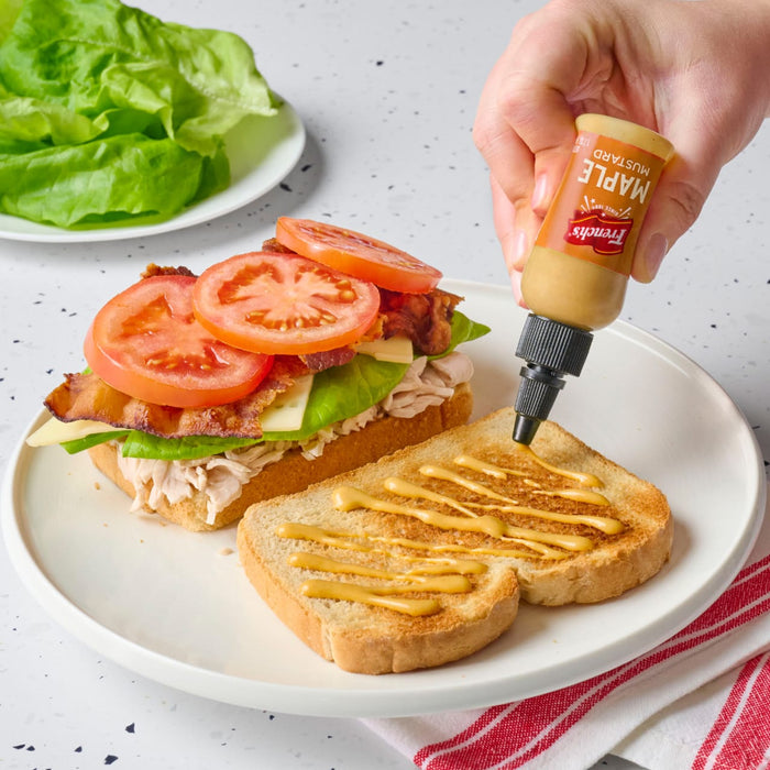 A hand squeezes French's Maple Mustard onto a sandwich with turkey, bacon, lettuce, and tomato.