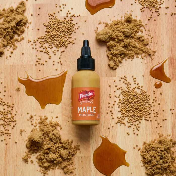 A bottle of French's Maple Mustard is surrounded by piles of brown sugar, mustard seeds, and maple syrup on a wooden surface.