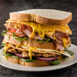 ['A close-up of a large sandwich with ham, chicken, cheese, lettuce, tomato, onion, and mustard.']