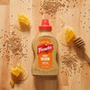 A bottle of Frenchs Brown Honey Dijon Mustard 12 Oz is centered on a wooden surface surrounded by scattered mustard seeds and pieces of honeycomb dripping with honey.