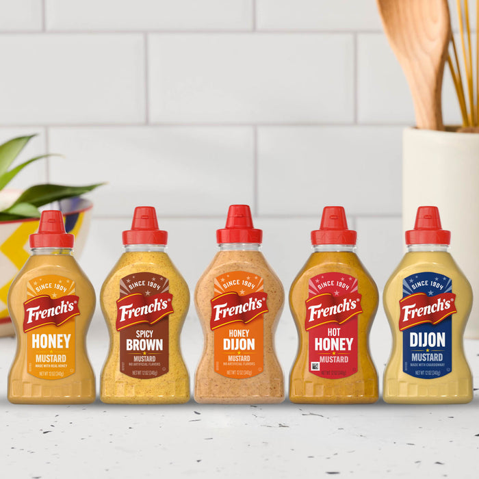 A lineup of five different varieties of Frenchs Brown Honey Dijon Mustard 12 Oz bottles including Honey, Spicy Brown, Honey Dijon, Hot Honey, and Dijon flavors displayed against a white tiled kitchen background.