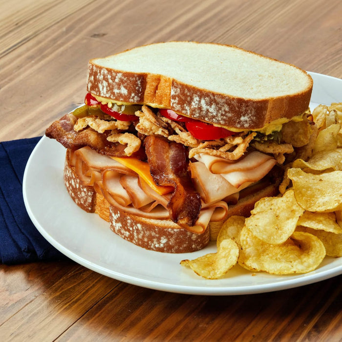 A hearty turkey and bacon club sandwich on thick sliced bread is layered with Frenchs Brown Honey Dijon Mustard 12 Oz and served with a side of crunchy potato chips.