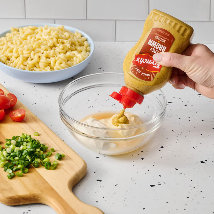 A hand squeezes a bottle of Frenchs Brown Spicy Brown Mustard 12 Oz into a glass bowl to create a creamy dressing for a pasta salad.