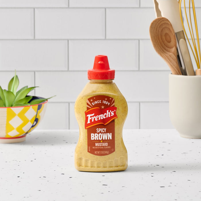 A bottle of Frenchs Brown Spicy Brown Mustard 12 Oz is centered on a white kitchen countertop, ready to be used in a recipe.