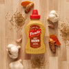 A bottle of Frenchs Brown Spicy Brown Mustard 12 Oz is artfully arranged on a wooden surface, surrounded by its key ingredients: whole garlic bulbs, mustard seeds, and fresh turmeric root.