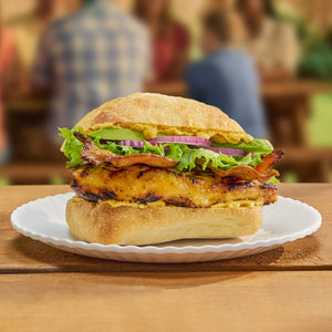 A grilled chicken sandwich with bacon, lettuce, avocado, and red onion is generously spread with Frenchs Brown Spicy Brown Mustard 12 Oz and served on a paper plate.