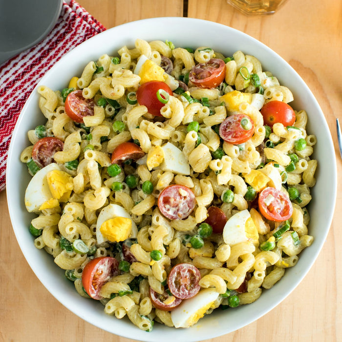 A bowl of creamy macaroni salad with hard-boiled eggs, peas, and tomatoes is shown, made with a flavorful dressing using Frenchs Brown Spicy Brown Mustard 12 Oz.