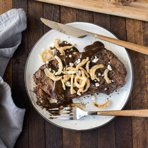 A steak with a brown sauce crispy onion rings and blue cheese crumbles on a white plate.