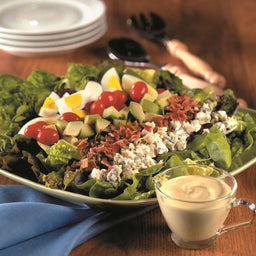A salad with bacon avocado blue cheese tomatoes and hard-boiled eggs is served with a creamy dressing.
