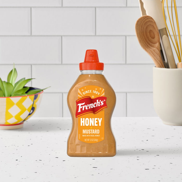 A bottle of Frenchs Brown Honey Mustard 12 Oz is centered on a white speckled countertop, with a bowl of greens and a utensil holder in the background.