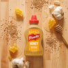 A bottle of Frenchs Brown Honey Mustard 12 Oz is placed on a wooden surface, surrounded by its key ingredients: mustard seeds, fresh garlic, and pieces of honeycomb with drizzled honey.