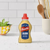 A bottle of Frenchs Beige Dijon Chardonnay Mustard 12 Oz is centered on a white speckled kitchen counter, with a white tiled backsplash and other kitchen items blurred in the background.