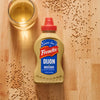 A bottle of Frenchs Beige Dijon Chardonnay Mustard 12 Oz is styled on a wooden cutting board with mustard seeds and a glass of white wine to highlight its ingredients.