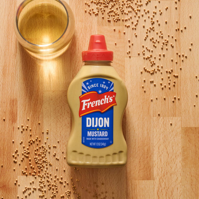 A bottle of Frenchs Beige Dijon Chardonnay Mustard 12 Oz is styled on a wooden cutting board with mustard seeds and a glass of white wine to highlight its ingredients.