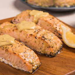 Several cooked salmon fillets are topped with a creamy sauce made from Frenchs Beige Dijon Chardonnay Mustard 12 Oz and served on a wooden board with a lemon wedge.