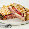 A delicious roast beef panini, cut in half to show the melted cheese, tomato, and red onion, is spread with Frenchs Beige Dijon Chardonnay Mustard 12 Oz and served with a side of potato chips.
