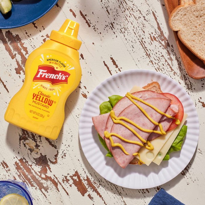 ["A bottle of French's Classic Yellow Mustard next to a sandwich with ham, cheese, tomato, and lettuce."]