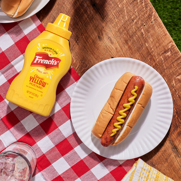 A bottle of French's Classic Yellow Mustard next to a hot dog on a white plate.